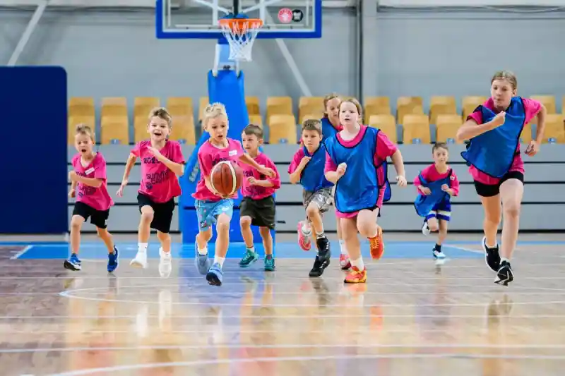 Bērni rozā un zilos treniņkreklos skrien pēc bumbas basketbola zālē